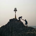 Silhouette of two people helping each other on a rocky hill with a trident at the peak.